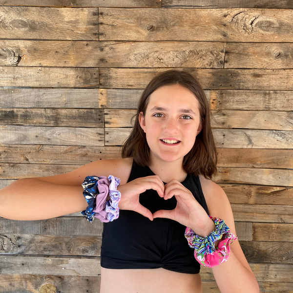 Close up picture of abstract navy blue scrunchie pictured with other swim scrunchies. Scrunchies are worn on the wrist of the owner and she is making a heart symbol with her hands. Pipevine Designs.