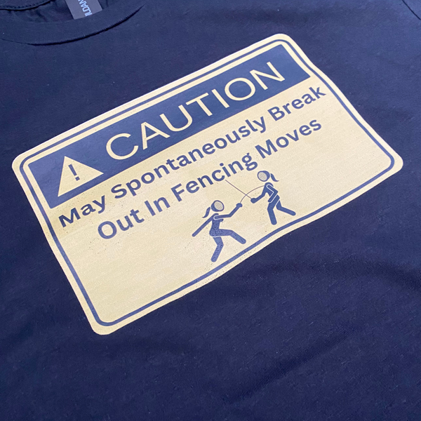 A closeup of a yellow warning sign featuring the words, "Caution. May spontaneously break out in fencing moves.", with two girl fencers demonstrating the danger. Black adult shirt.