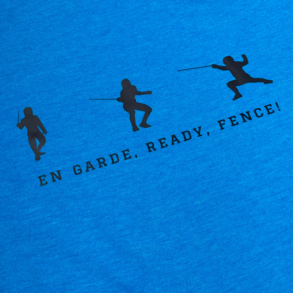 A closeup of a sapphire blue shirt, featuring three silhouetted fencers, each one in a different fencing stance. The positions include the classic stance, a slightly lower and readier stance, and the full extended lunge. The words, "En Garde, Ready, Fence!" hover beneath, narrating the scenes above. Adult shirt.