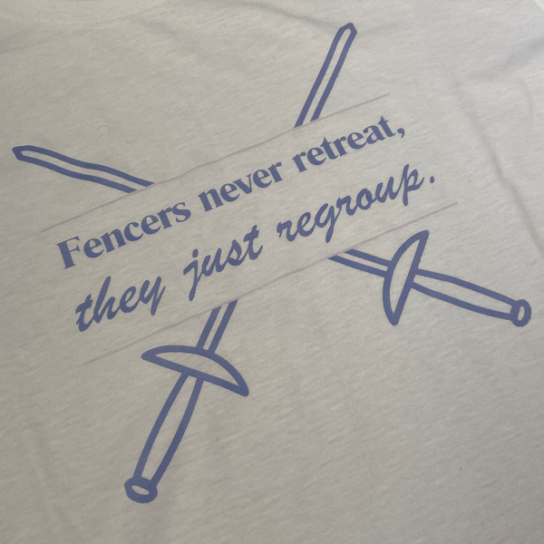 a closeup of the purple words, "Fencers never retreat, they just regroup." are bordered by light pink lines, and two crisp, purple sword outlines, the same color as the words, crisscross behind. Adult white shirt.