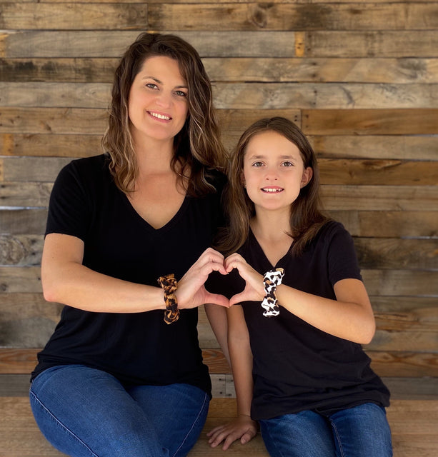 A mother and daughter duo smiling at the camera while making a heart symbol with their hands. Both are wearing colorful and stylish scrunchies in a leopard print, adding a pop of color to the image. The two are shown in a friendly and loving pose, creating a heartwarming and positive moment. 