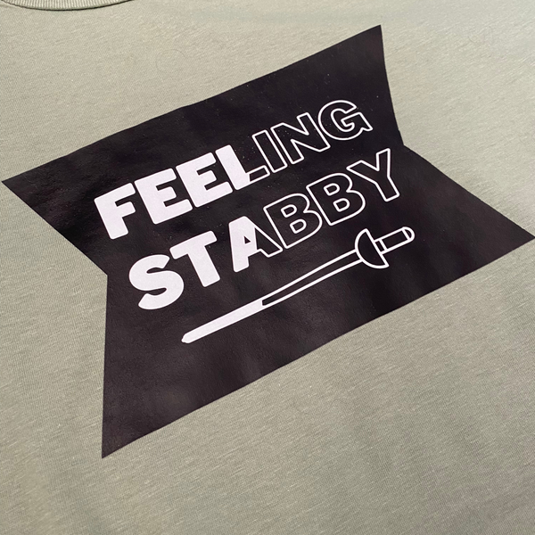 Closeup of a black emblem, featuring the humorous words, "Feeling Stabby", with a sword below. The words and sword are split diagonally, changing them from having white filling to just an outline, making the whole shirt more eye-catching. A pink adult shirt.